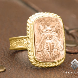 May include: A gold ring with a detailed design featuring a raised, textured image of a moth. The ring has a square-shaped setting with a rope-like border.