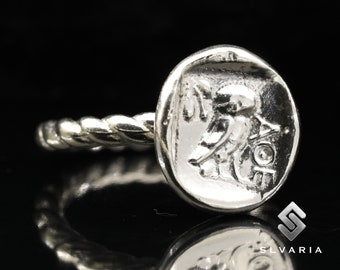 May include: Silver ring with a twisted band and a round, coin-shaped top featuring a detailed engraving of a winged figure.
