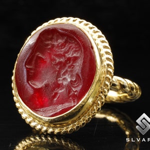 May include: A gold ring with a red cameo featuring a profile of a person with a crown. The ring has a rope-like design around the edge.