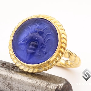 May include: A gold ring with a blue glass cameo featuring a detailed engraved design of a bee. The ring has a rope-like band.