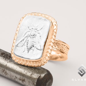 May include: A gold-toned ring with a rectangular face featuring a detailed bee design. The ring has a rope-like border around the face and ornate detailing on the band. The bee is silver-colored, contrasting with the gold of the ring.