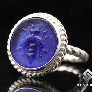 May include: A silver ring with a blue glass cameo featuring a detailed bee design. The ring has a rope-like band.