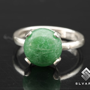 Könnte beinhalten: Ein silberner Ring mit einem grünen Edelstein, der in einer Krappenfassung sitzt. Der Ring befindet sich auf einem schwarzen Hintergrund.