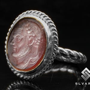 May include: A silver ring with a red cameo featuring a carved profile of a person's head. The ring has a rope-like band.