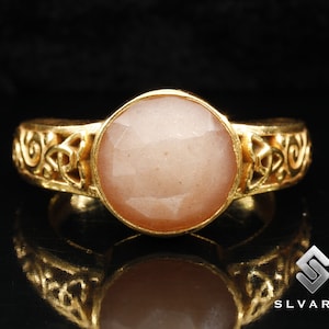 May include: A gold ring with a large, round, light pink gemstone set in a Celtic knot design.