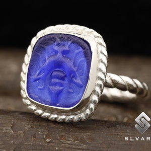 May include: A silver ring with a blue glass cameo featuring a detailed design of a mythical creature. The ring has a rope-like band.