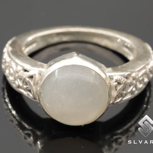 Op de afbeelding: Een zilveren ring met een grote, ronde, bleke edelsteen. De band heeft een ingewikkeld, zich herhalend patroon. Het ontwerp van de ring is eenvoudig maar elegant, geschikt voor dagelijks gebruik of speciale gelegenheden.