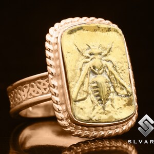 May include: A gold ring with a Celtic knot design on the band and a square bezel featuring a raised image of a bee. The bezel is surrounded by a rope-like border.