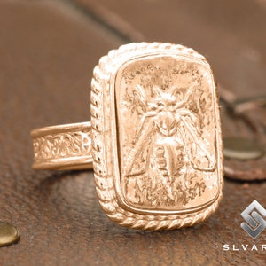 May include: A gold ring with a detailed design of a moth on the face. The ring has a rope-like border around the face.