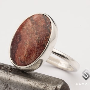 Può includere: Un anello d'argento con una grande pietra ovale di colore rosso e marrone incastonata al centro. L'anello è appoggiato su una superficie metallica con il numero 925 stampato.