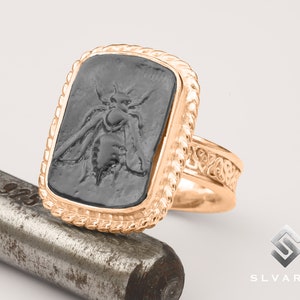 May include: A gold ring with a black rectangular stone featuring an engraved bee design. The ring has a rope-like border around the stone and a detailed band.
