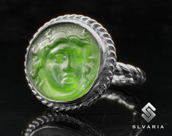 May include: A silver ring with a green glass cameo featuring a face with a detailed design. The ring has a rope-like band.