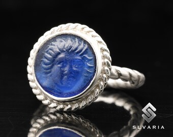 May include: A silver ring with a blue glass cameo featuring a face with a detailed hairstyle. The ring has a rope-like design around the edge of the cameo.