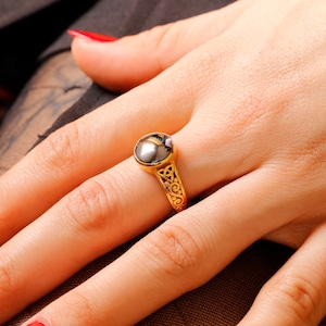 May include: A gold ring with a black oval gemstone and an intricate design. The ring is on a person's finger.