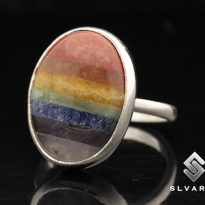 May include: A silver ring with a rainbow-colored oval gemstone. The gemstone is made of seven different colors of stones, including red, orange, yellow, green, blue, indigo, and violet.