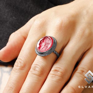 May include: A silver ring with a red cameo featuring a woman's profile. The cameo is set in a silver bezel with a rope-like design.