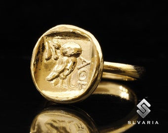 May include: A gold ring with a raised design of a winged figure holding a staff. The ring is set against a black background.