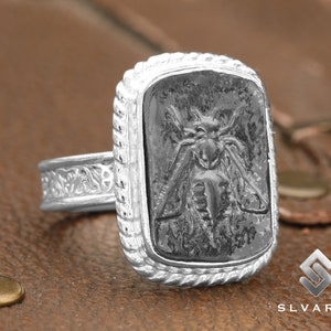 May include: A silver ring with a detailed, ornate band and a rectangular black stone featuring a carved insect design.