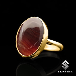 May include: A gold ring with a large oval-shaped red gemstone. The gemstone has a striated pattern.