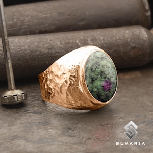 May include: A gold ring with a hammered finish and a green oval gemstone set in the centre. The gemstone has a pink hue and a speckled pattern.
