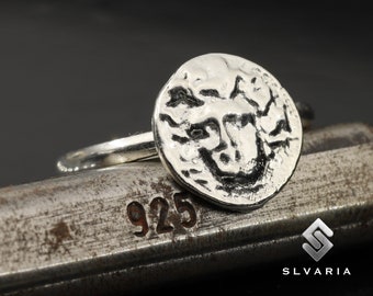 May include: A silver ring with a textured, round face featuring a detailed design of a mythical creature. The ring is resting on a metal surface with the number 925 stamped on it.