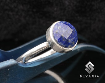 May include: A silver ring with a round, faceted, dark blue lapis lazuli gemstone. The ring is set on a blue surface.