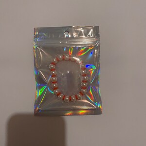 May include: A pearl bead bracelet with orange spacers is displayed inside a holographic, iridescent plastic bag. The bracelet is made of round, white beads. A simple, elegant piece of jewellery.