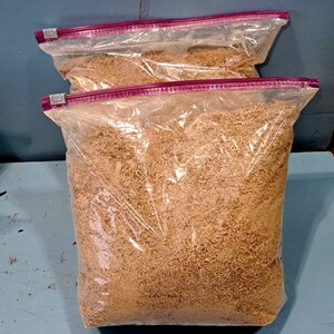 Puede incluir: Dos bolsas de pl&aacute;stico transparente llenas de virutas de madera de color marr&oacute;n claro. Las bolsas tienen un cierre de cremallera rosa y est&aacute;n selladas. Las bolsas est&aacute;n sobre una superficie azul.
