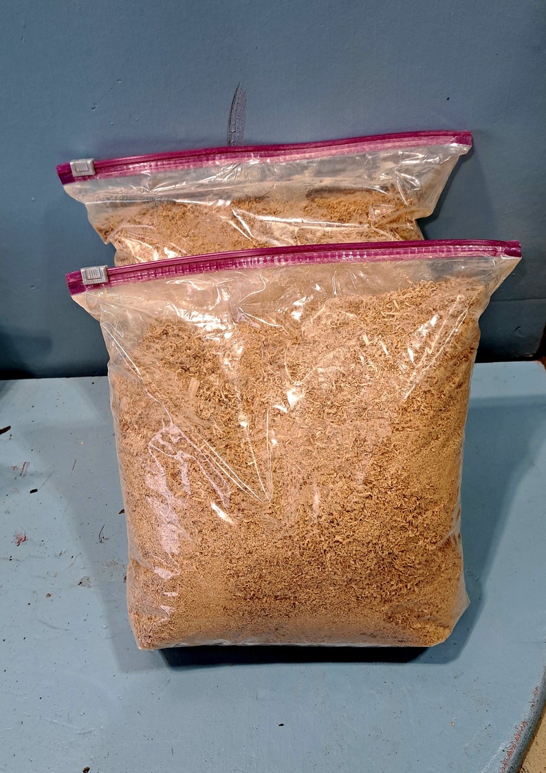 Puede incluir: Dos bolsas de pl&aacute;stico transparente llenas de virutas de madera de color marr&oacute;n claro. Las bolsas tienen un cierre de cremallera rosa y est&aacute;n selladas. Las bolsas est&aacute;n sobre una superficie azul.