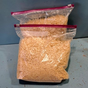 Puede incluir: Dos bolsas de pl&aacute;stico transparente llenas de virutas de madera de color marr&oacute;n claro. Las bolsas tienen un cierre de cremallera rojo y est&aacute;n apiladas. El fondo es una superficie azul-verdosa.