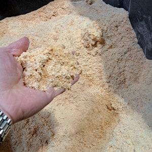 Puede incluir: Primer plano de una mano que sostiene un pu&ntilde;ado de virutas de madera de color marr&oacute;n claro, con una textura &aacute;spera. Las virutas est&aacute;n en un gran recipiente negro, lleno de m&aacute;s virutas.