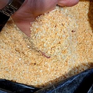 Puede incluir: Primer plano de una mano sosteniendo un pu&ntilde;ado de virutas de madera de color marr&oacute;n claro. Las virutas son gruesas y texturizadas, con una mezcla de tonos claros y oscuros. Se puede ver una peque&ntilde;a lombriz de tierra rosa entre las virutas. Las virutas est&aacute;n contenidas en una bolsa de pl&aacute;stico negra.