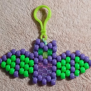 Witch's Brew aethetic Kandi Bat with a purple body and lime green wings, UV reactive transparent purple belly, UV reactive transparent yellow nose, and UV reactive transparent green eyes, done on rainbow stretchy cord and finished with a lime green hook.