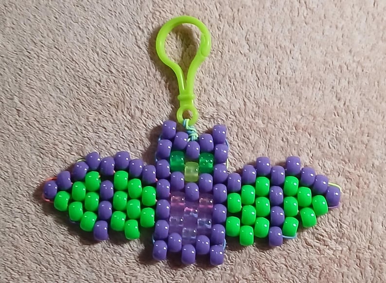 Witch's Brew aethetic Kandi Bat with a purple body and lime green wings, UV reactive transparent purple belly, UV reactive transparent yellow nose, and UV reactive transparent green eyes, done on rainbow stretchy cord and finished with a lime green hook.