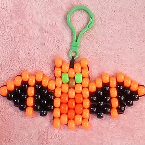 Jack o'lantern aethetic Kandi Bat with a bright orange body, black wings, orange belly, brown nose, green eyes, done on black stretchy cord and finished with a green hook.