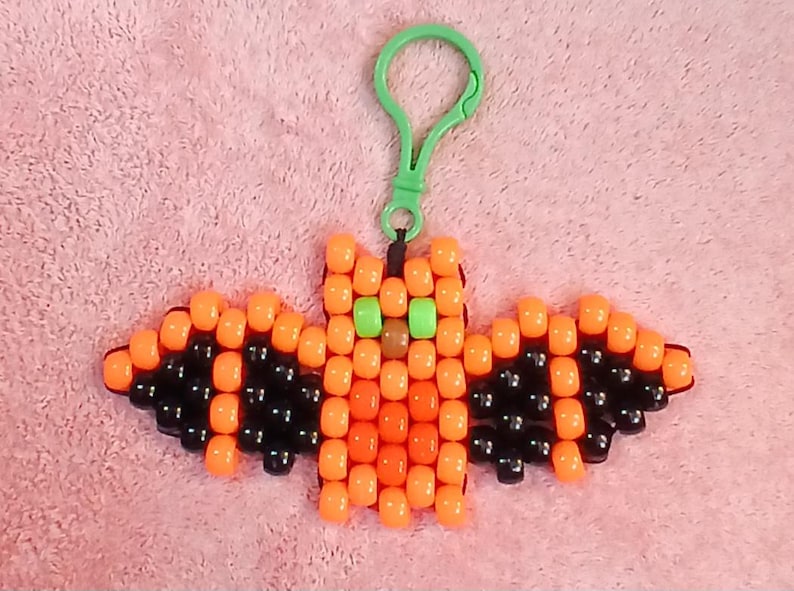 Jack o'lantern aethetic Kandi Bat with a bright orange body, black wings, orange belly, brown nose, green eyes, done on black stretchy cord and finished with a green hook.