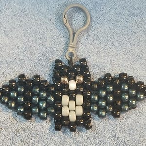 Black aethetic Kandi Bat with a black body, gunmetal wings, grey belly, white nose, and silver eyes, done on black stretchy cord and finished with a grey hook.
