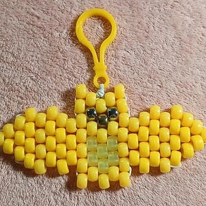 Yellow aethetic Kandi Bat with semiopaque body and opaque wings, UV reactive transparent yellow belly, bronze nose, and gold eyes, done on white stretchy cord and finished with a matching yellow hook.