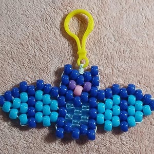 Blue aethetic Kandi Bat with a dark blue body and light blue wings, UV reactive transparent blue belly, light pink nose, and purple eyes, done on white stretchy cord and finished with a yellow hook.