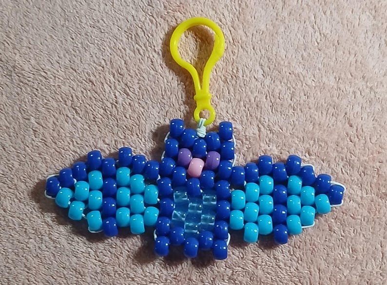 Blue aethetic Kandi Bat with a dark blue body and light blue wings, UV reactive transparent blue belly, light pink nose, and purple eyes, done on white stretchy cord and finished with a yellow hook.