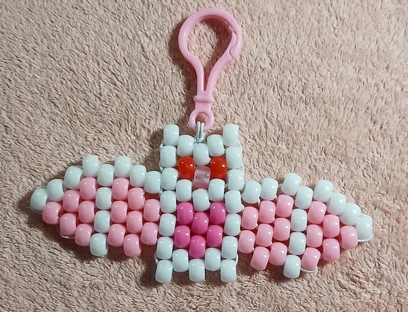 Coquette aethetic Kandi Bat with a white body and light pink wings, pink belly, UV reactive transparent light pink nose, and red eyes, done on white stretchy cord and finished with a matching light pink hook.