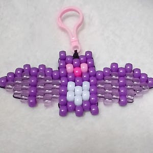 Purple aesthetic Kandi Bat with purple body, UV reactive transparent wings, white stomach, light pink eyes, and a pink nose, done on black stretchy cord and finished with a light pink hook