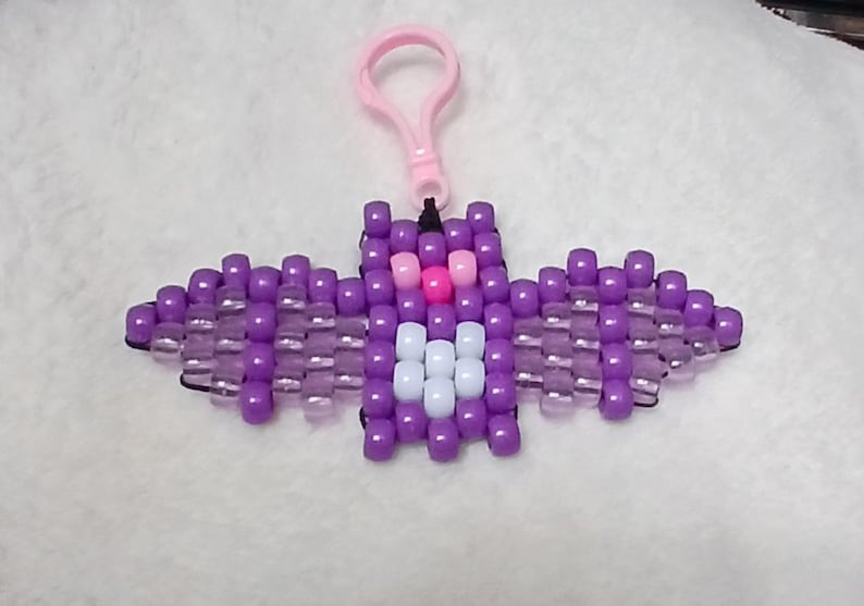 Purple aesthetic Kandi Bat with purple body, UV reactive transparent wings, white stomach, light pink eyes, and a pink nose, done on black stretchy cord and finished with a light pink hook