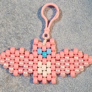 Light Pink aethetic Kandi Bat with a light pink body, light pink pearl wings, white belly, light blue nose, and pastel blue eyes, done on black stretchy cord and finished with a light pink hook.