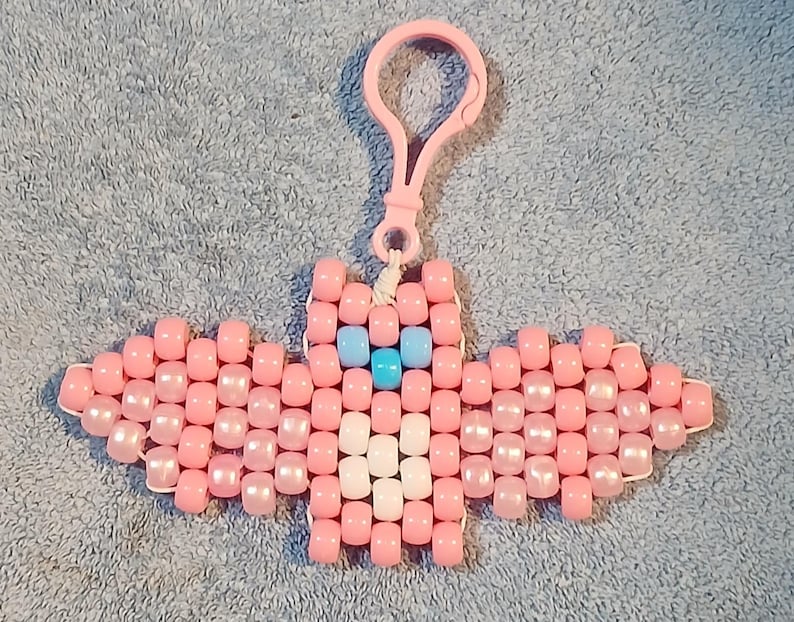 Light Pink aethetic Kandi Bat with a light pink body, light pink pearl wings, white belly, light blue nose, and pastel blue eyes, done on black stretchy cord and finished with a light pink hook.