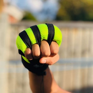 Op de afbeelding: Een close-up van een hand die een vingerloze trainingshandschoen draagt in zwart en neon groen. De handschoen heeft een mesh-ontwerp en wordt vastgezet met een zwarte band.