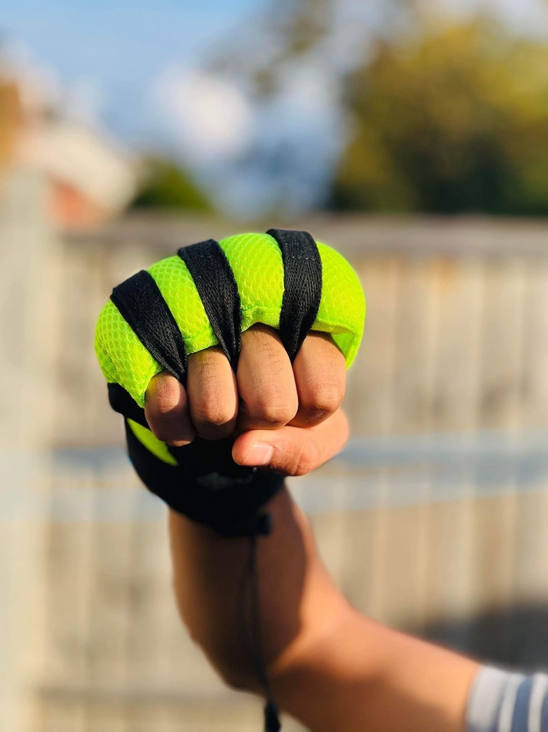 Op de afbeelding: Een close-up van een hand die een vingerloze trainingshandschoen draagt in zwart en neon groen. De handschoen heeft een mesh-ontwerp en wordt vastgezet met een zwarte band.
