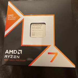 Op de afbeelding: Een zwarte doos met oranje en zilveren accenten, met de AMD Ryzen 7 9000 Series Processor. De doos heeft het AMD Ryzen logo en het nummer 7 in oranje. De processor zelf is zichtbaar in een vierkante uitsparing.