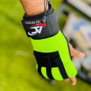 Op de afbeelding: Groene en zwarte gewichthefgloves met een polsband. De band heeft de tekst "AC" en "CES SPORTS" in rood.