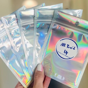 May include: A stack of iridescent silver resealable bags with a white circle sticker on the front that says "All Tea'd Up" with blue text and "Follow us on Facebook" in smaller blue text.